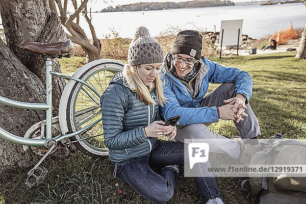 Young couple sitting on grass and using smart phone during break from bike ride  Portland  Maine  USA