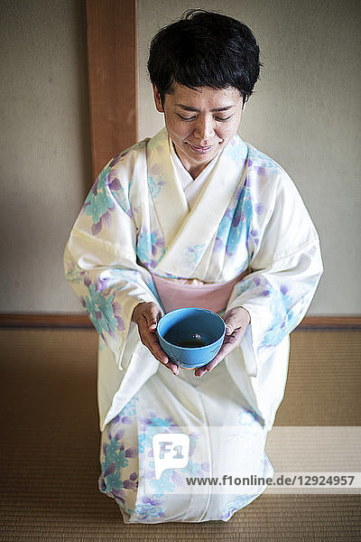 Japanische Frau in traditionellem weißen Kimono mit blauem Blumenmuster  die während der Teezeremonie auf dem Boden kniet und eine blaue Teeschale hält.
