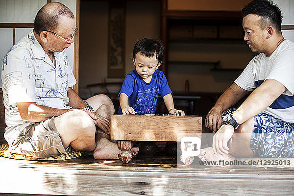 Zwei japanische Männer und ein kleiner Junge sitzen auf dem Boden auf der Veranda eines traditionellen japanischen Hauses und spielen Go.