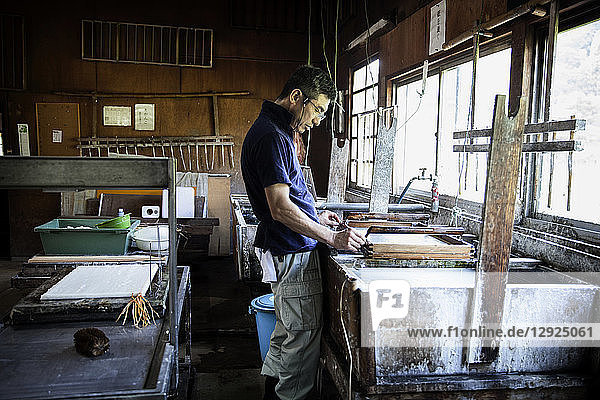 Japanischer Mann in einer Werkstatt  der einen Holzrahmen mit gepresstem Zellstoff hält und traditionelles Washi-Papier herstellt.