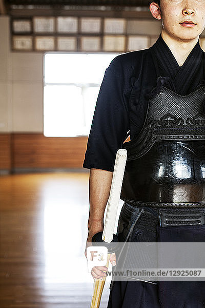 Männlicher japanischer Kendo-Kämpfer  der in einer Turnhalle steht und ein Holzschwert hält.