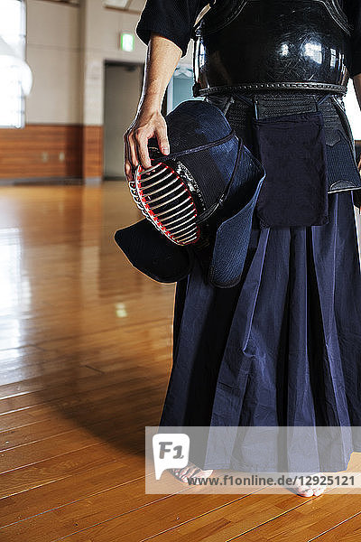 Male Japanese Kendo fighter standing in a gym  holding Kendo mask.