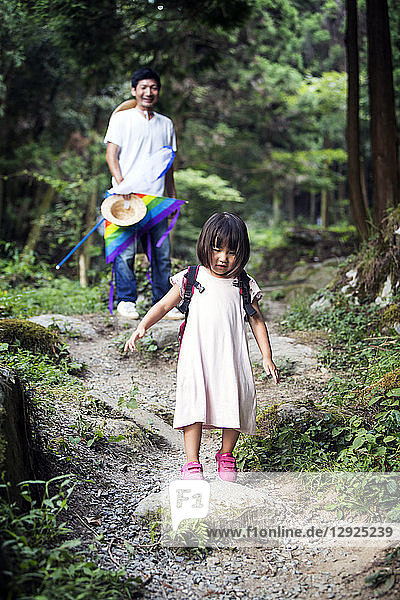 Im Wald stehendes japanisches Mädchen in hellrosa Sonnenkleid und mit Rucksack  im Hintergrund ein Mann.