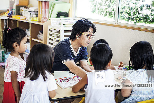 Männlicher Lehrer im Gespräch mit einer Gruppe von Kindern an einem Tisch in einer japanischen Vorschule.