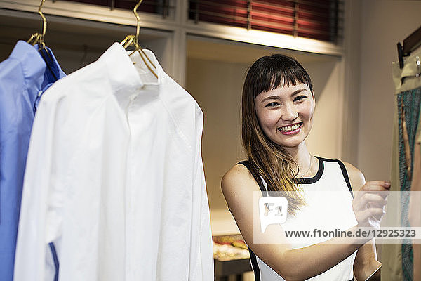 Lächelnde japanische Verkäuferin steht im Bekleidungsgeschäft und schaut auf das Hemd.