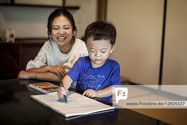 Lächelnde japanische Frau und kleiner Junge sitzen an einem Tisch und zeichnen mit Farbstiften auf weißes Papier.