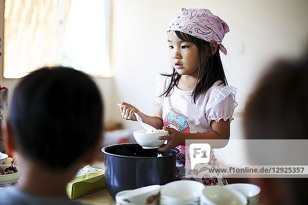 Girl wearing headscarf standing at a table in a Japanese preschool  serving lunch to children.