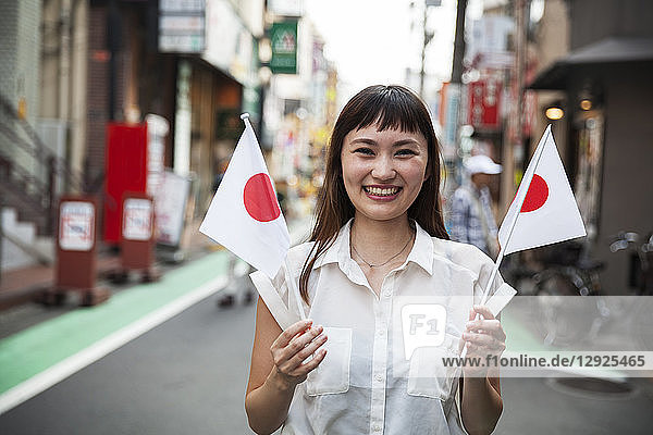 Lächelnde japanische Frau mit langen braunen Haaren und weißer  kurzärmeliger Bluse  die in einer Straße steht und eine kleine japanische Flagge hält.