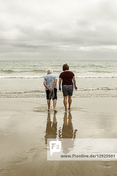 Rear view of a grey haired elderly woman and a younger woman paddling with shoes off in shallow waves on a sandy beach.
