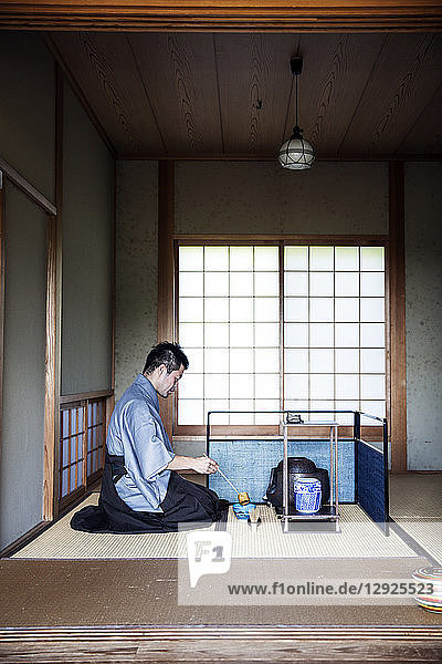 Japanese man wearing traditional kimono kneeling on floor using a Hishaku a bamboo ladle during a tea ceremony.