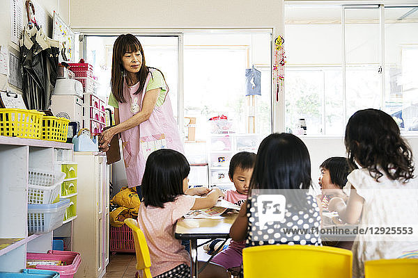 Eine junge Lehrerin und eine Gruppe von Kindern in einer japanischen Vorschule an einem Tisch sitzend.