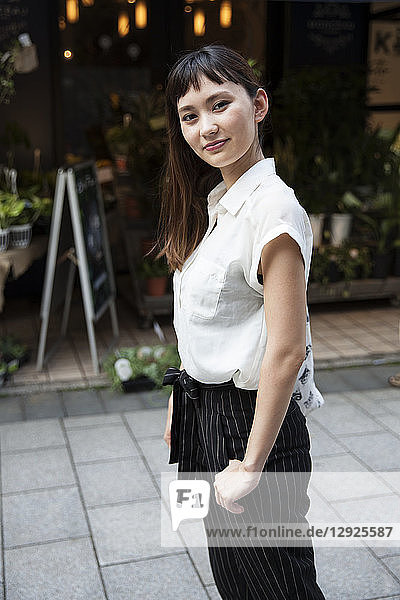 Japanerin mit langen braunen Haaren und weißer  kurzärmeliger Bluse  steht in einer Straße und lächelt in die Kamera.