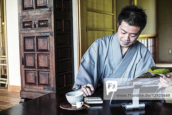 Japanischer Mann in Kimono  der in einem traditionellen japanischen Haus auf dem Boden sitzend einen Taschenrechner und ein digitales Tablett benutzt.