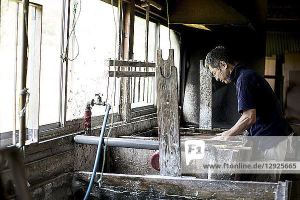 Japanischer Mann in einer Werkstatt  der sich über einen Bottich mit Flüssigkeit beugt  dem traditionellen Washi-Papierherstellungsverfahren.