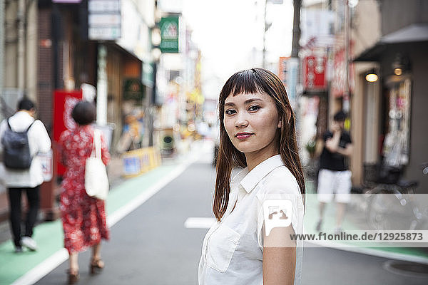 Smiling Japanese woman with long brown hair wearing white short-sleeved blouse standing in a street.