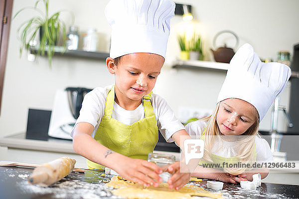 Two young kids happy childrens boy and girl family with apron and chef hat preparing funny cookies in kitchen at home.