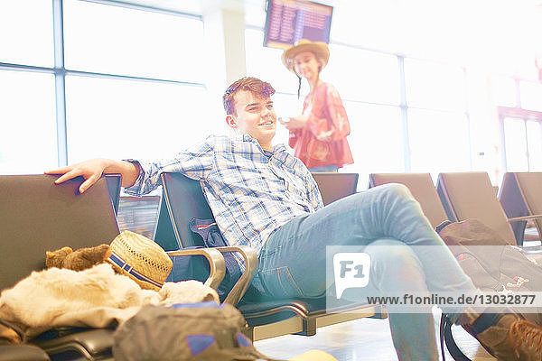 Junger Mann sitzt am Flughafen  Rucksack neben ihm  junge Frau steht neben ihm und schaut zu ihm