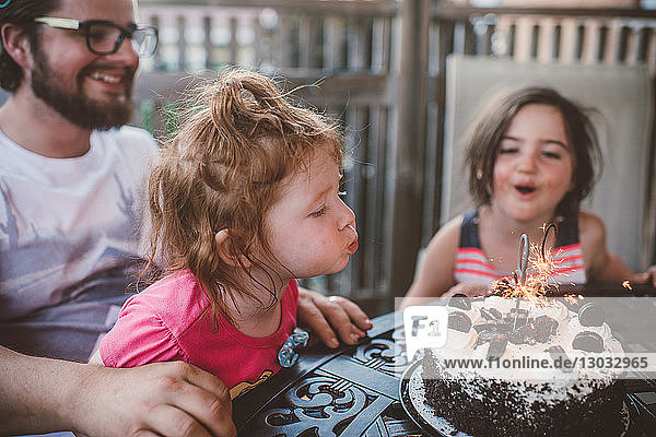 Female toddler with sister and father blowing out sparklers on celebration cake at patio table
