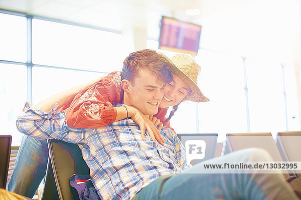 Junger Mann sitzt am Flughafen  junge Frau streckt sich über den Sitz  um ihn zu umarmen