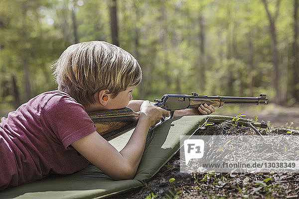 Boy aiming rifle while lying on mat in forest