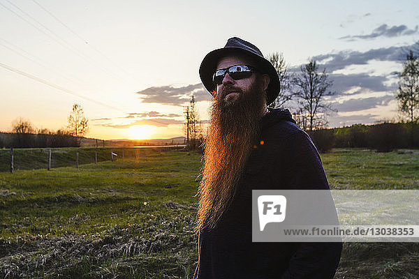Bearded man wearing hat and sunglasses looking away while standing on field against sky during sunset