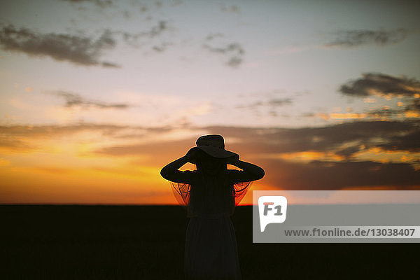Silhouettenmädchen mit Hut  das bei Sonnenuntergang am Himmel steht