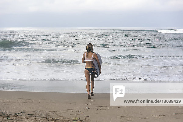 Rückansicht einer Frau  die ein Surfbrett trägt  während sie am Strand auf das Meer zuläuft