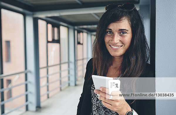 Porträt einer lächelnden Frau  die ein Mobiltelefon benutzt  während sie auf einer Fußgängerbrücke steht