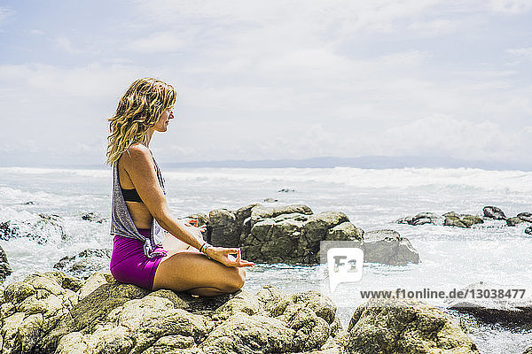 Seitenansicht einer mittleren erwachsenen Frau  die in Lotus-Position auf Felsen am Strand meditiert