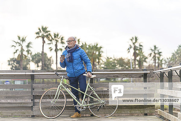 Mann hält Kaffee in der Hand  während er mit dem Fahrrad am Zaun des Parks steht
