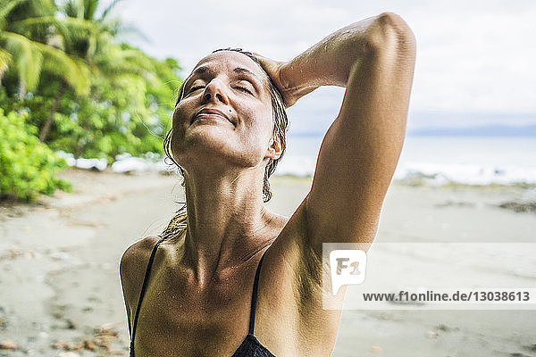 Entspannte Frau im mittleren Erwachsenenalter steht mit der Hand im nassen Haar am Strand