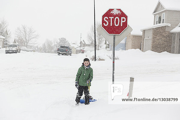 Teenager-Junge in voller Länge mit Schlitten  der im Winter auf schneebedeckter Straße mit Stoppschild steht