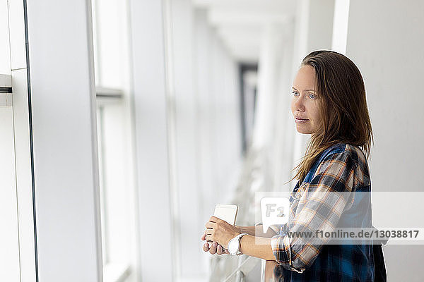 Nachdenkliche Frau schaut durch Fenster  während sie am Geländer im Flughafen steht