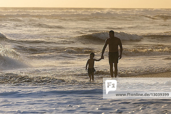 Vater ohne Shirt mit Sohn bei Sonnenuntergang am Strand spazieren