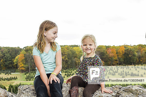 Portrait of smiling girl sitting by sister on rock against clear sky