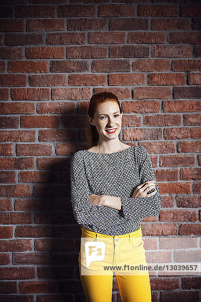 Portrait of happy businesswoman standing arms crossed against brick wall