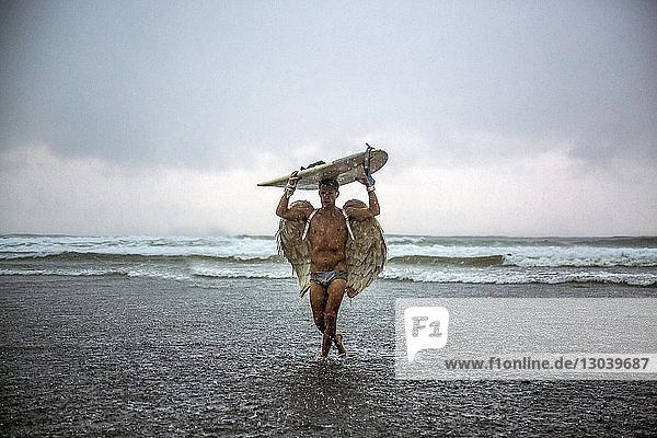 Man wearing angel wings carrying surfboard while walking in sea against cloudy sky