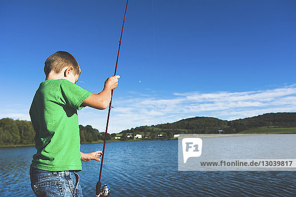 Side view of boy looking at fishing rod while standing against lake
