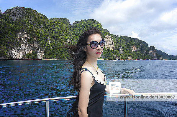 Seitenansicht einer jungen Frau mit Sonnenbrille  die an einer Reling auf einem Boot im Meer vor Bergen steht