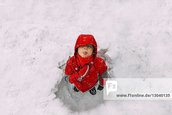 Hochwinkelaufnahme eines verspielten Jungen  der im Schnee stehend Schneeflocken auf der Zunge fängt