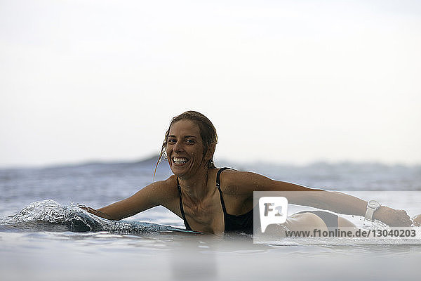 Portrait of happy woman lying on surfboard at beach against sky