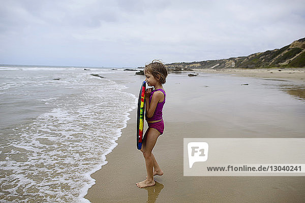 Side view of girl holding surfboard while standing at beach on Crystal Cove State Park against sky