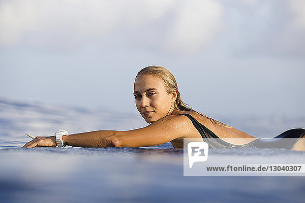 Portrait of confident woman lying on surfboard at beach against sky
