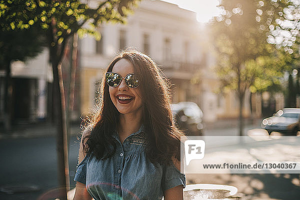 Fröhliche junge Frau mit Sonnenbrille  die am sonnigen Tag auf dem Bürgersteig in der Stadt steht