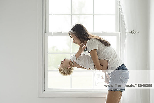 Side view of mother carrying son while playing against window at home
