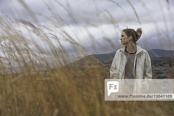 Thoughtful woman listening music while standing on grassy field against cloudy sky