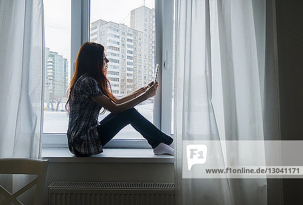 Side view of teenage girl using tablet computer while sitting by window at home