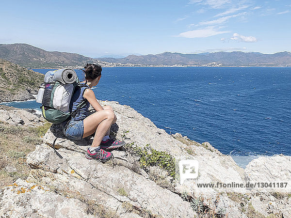 Wanderin mit Rucksack mit Blick auf die Aussicht  während sie bei Sonnenschein auf Felsen am Meer vor Bergen sitzt