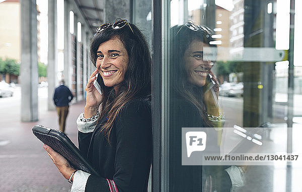 Porträt einer glücklichen Frau  die am Fenster stehend mit dem Handy telefoniert