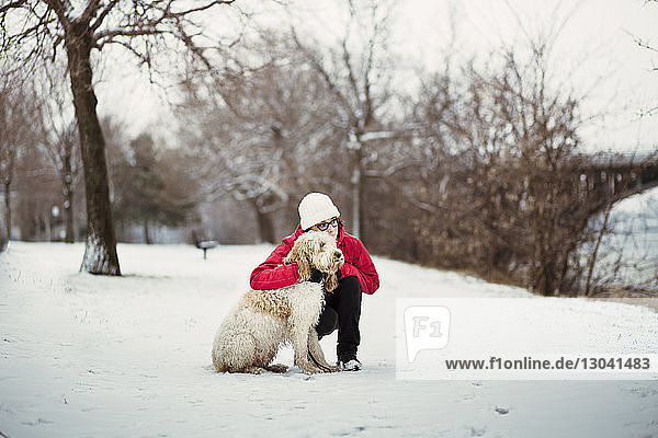 Woman with dog looking away while crouching on snow covered field
